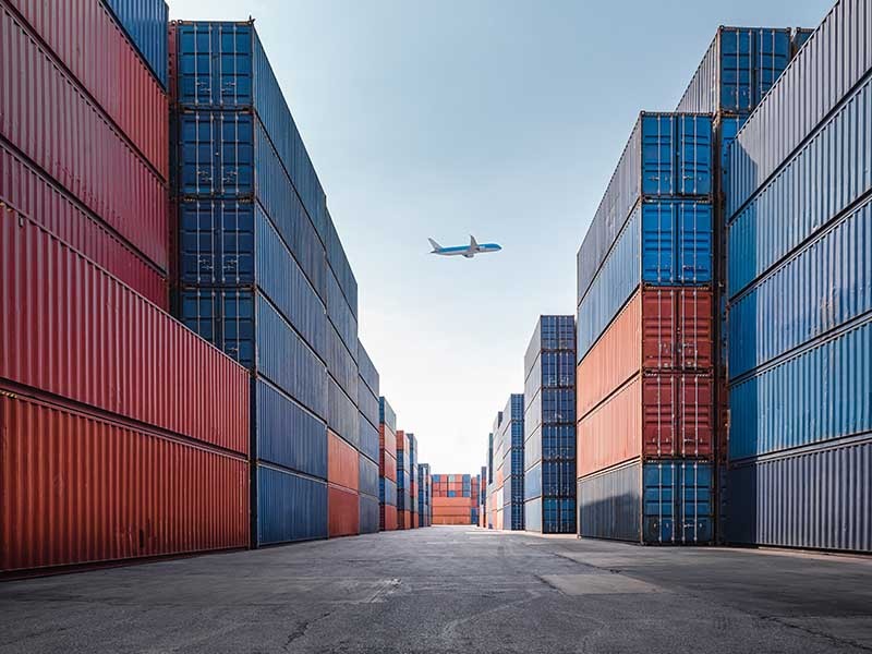 Airplane passing above huge shipping containers