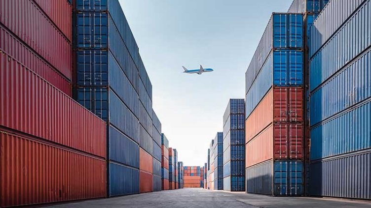 Airplane passing above huge shipping containers