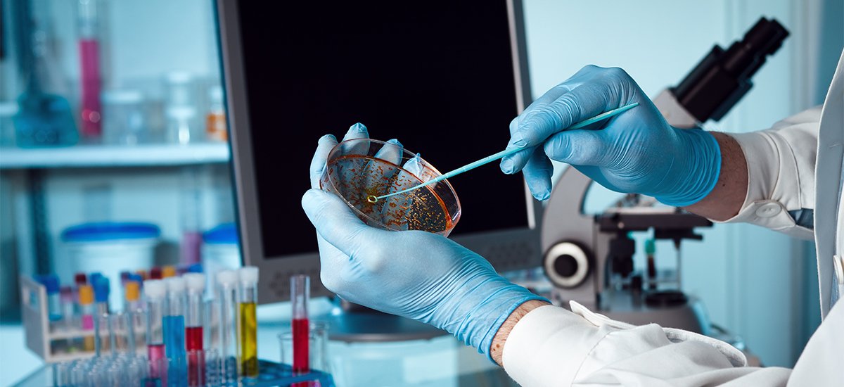 A scientist holding a petri dish in the lab with a monitor and microscope in background.