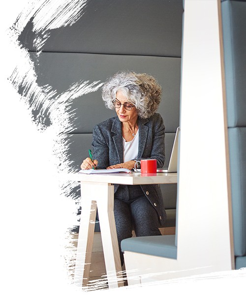 A lady sitting an writing on paper(cup of coffee and laptop in front of her)