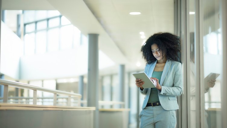 a young female businesswoman stands in the corridor  of a modern business interior looking at her digital tablet . The lighting is bright and there is a glow from the tablet device on her face. She is wearing a smart grey business suit .